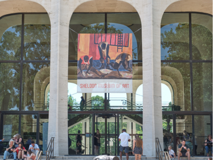 Modern building facade with large glass arches, steps, and art banner; people sitting and walking.