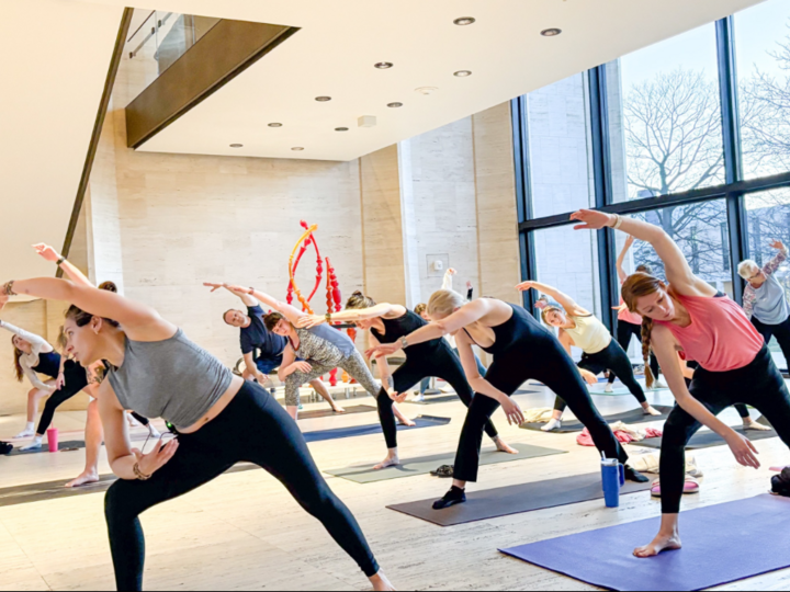 Yoga class in a bright studio with large windows, participants stretching in a side bend pose.